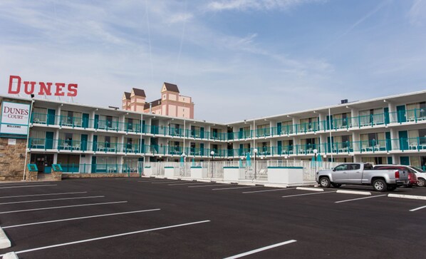 Un motel de varios pisos con un letrero que dice "DUNES COURT" y un estacionamiento al frente.