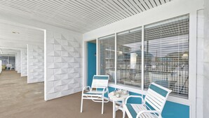 A covered outdoor seating area with white metal chairs and a table, white walls with geometric patterns, and large windows with blinds. - Dunes Court (Ocean City)