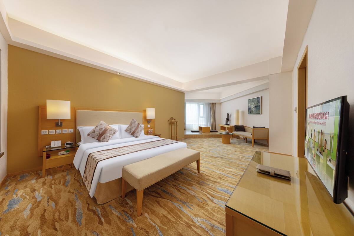 Een hotelkamer met een groot bed, nachtkastjes, een bankje, een televisie en een vloerbedekking.