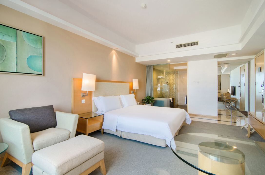Une chambre d’hôtel comprenant un lit, un coin salon avec un fauteuil et un pouf, une table basse en verre et un tableau au mur.