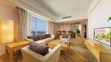Habitación de hotel moderna con sofá, sillón, mesa de centro, televisor y vistas a la ciudad.