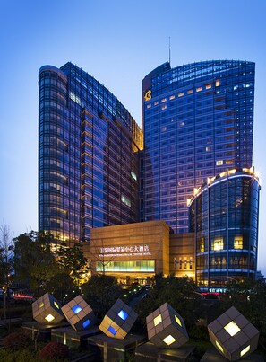 A modern hotel with a prominent glass facade, a central entrance with illuminated signage, and a distinctive cube-shaped structure in the foreground. - Fuyang International Trade Center Hotel (Hangzhou)