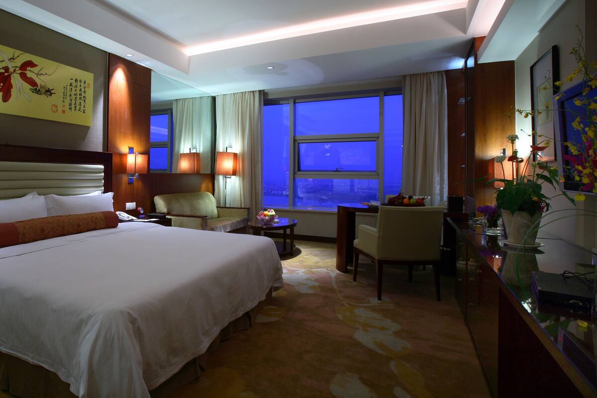 Une chambre d’hôtel avec un grand lit, un bureau, une chaise et une vue sur la ville la nuit.