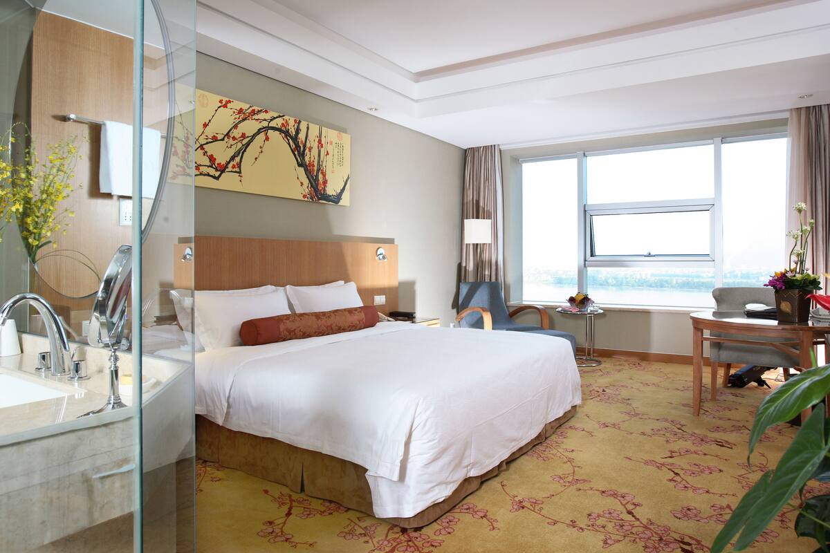 Une chambre d’hôtel avec un grand lit, un bureau, une chaise, une télévision, une fenêtre donnant sur un beau paysage et un tableau au mur.