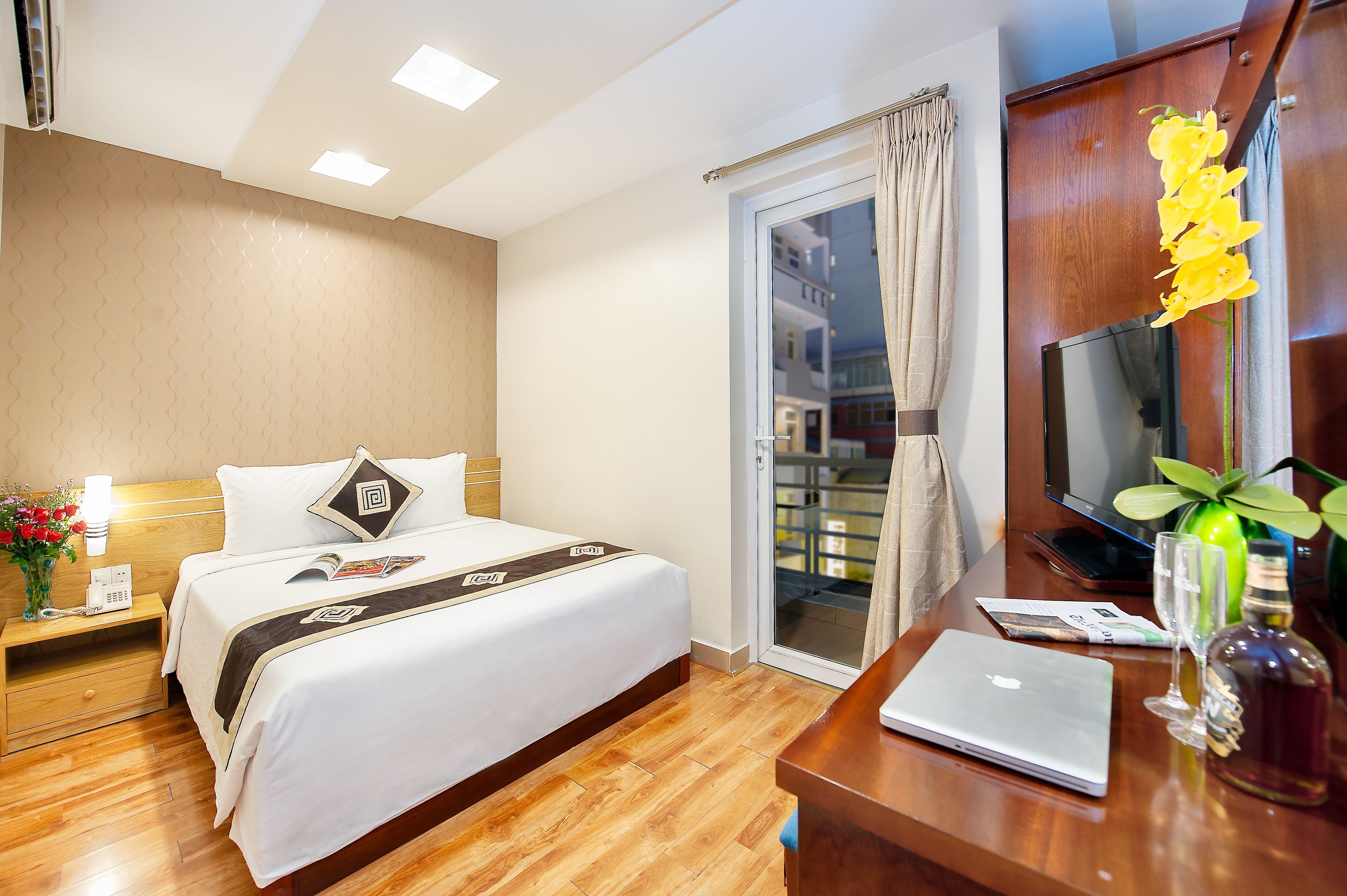 A hotel room with a bed, bedside table, a laptop, a bottle, and a plant.