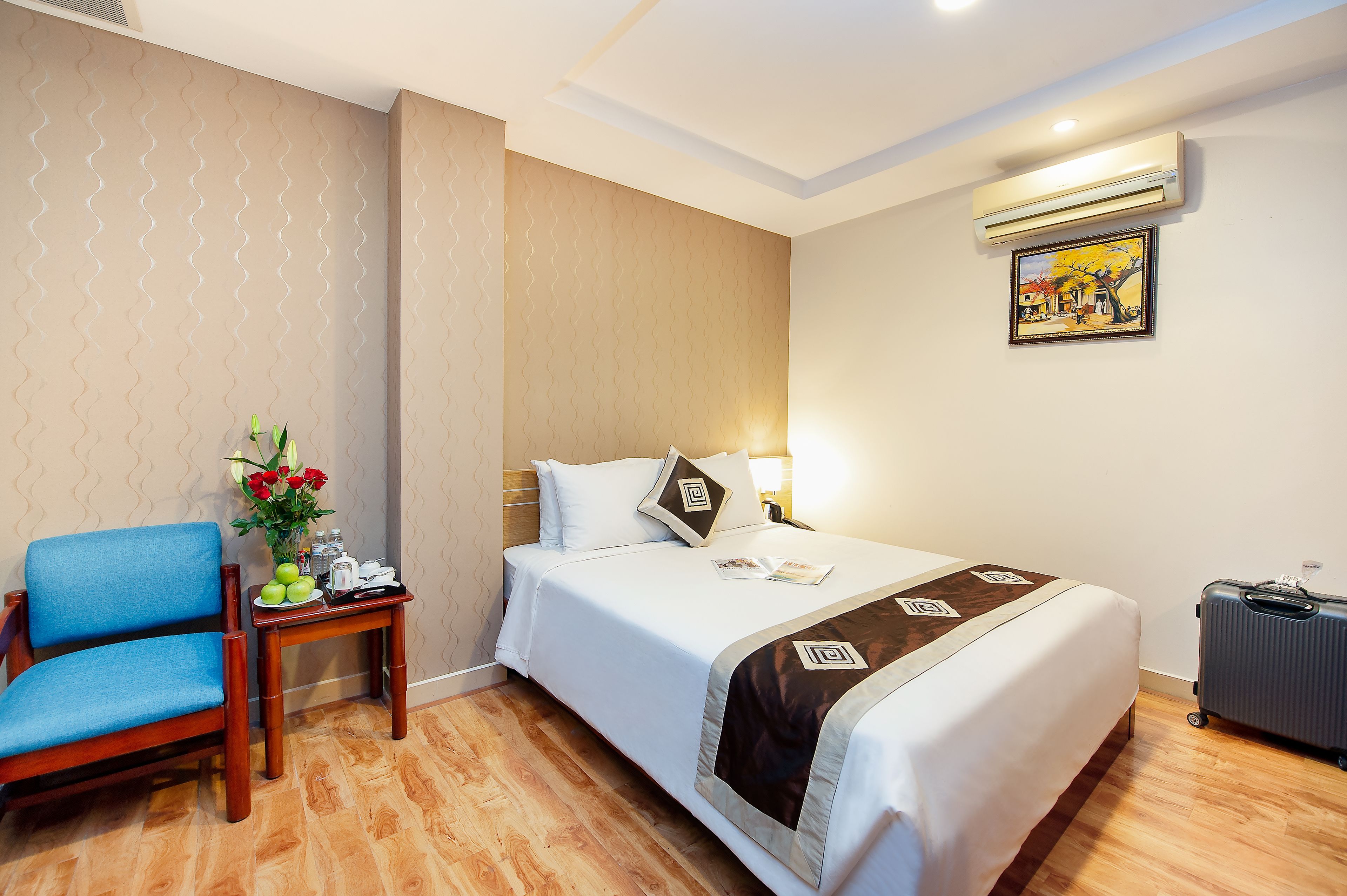 A hotel room with a bed, a blue chair, a small table with a vase of flowers, and a suitcase.