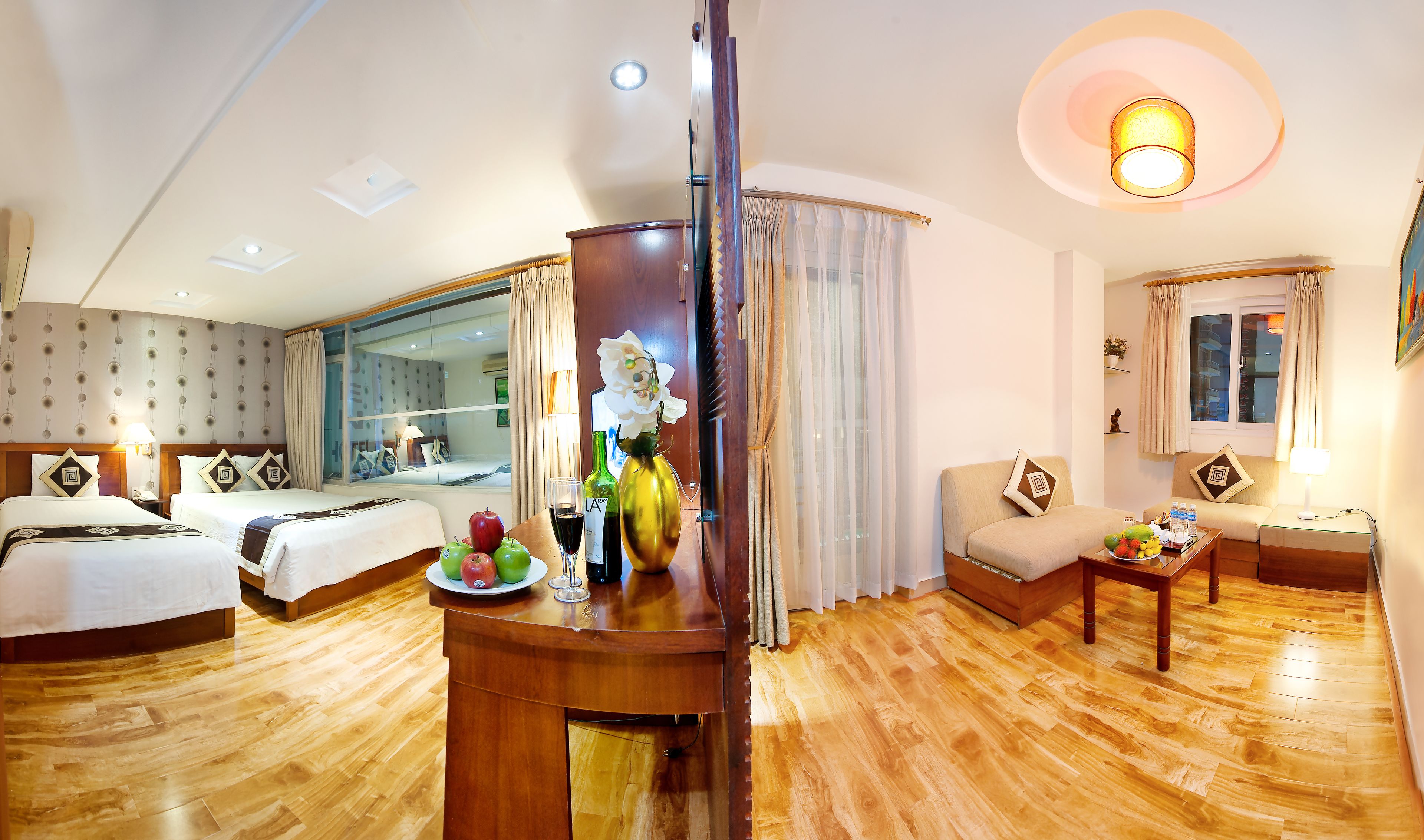 Une chambre d’hôtel avec deux lits, un canapé, une table à manger avec des fruits et du vin, et un miroir offrant une vue sur la ville.
