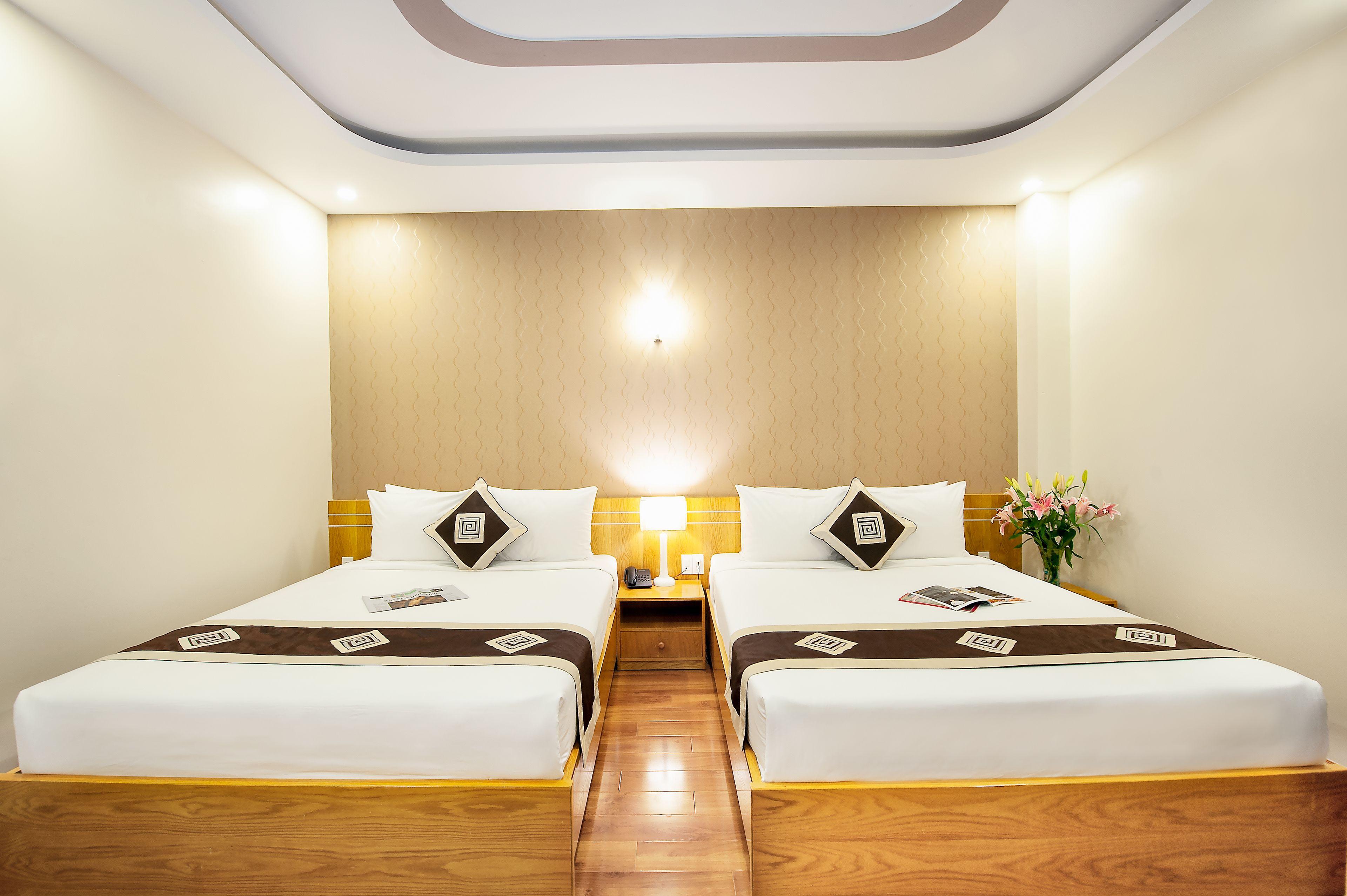 Habitación de hotel con dos camas individuales, piso de madera, mesita de noche con lámpara y un jarrón con flores.