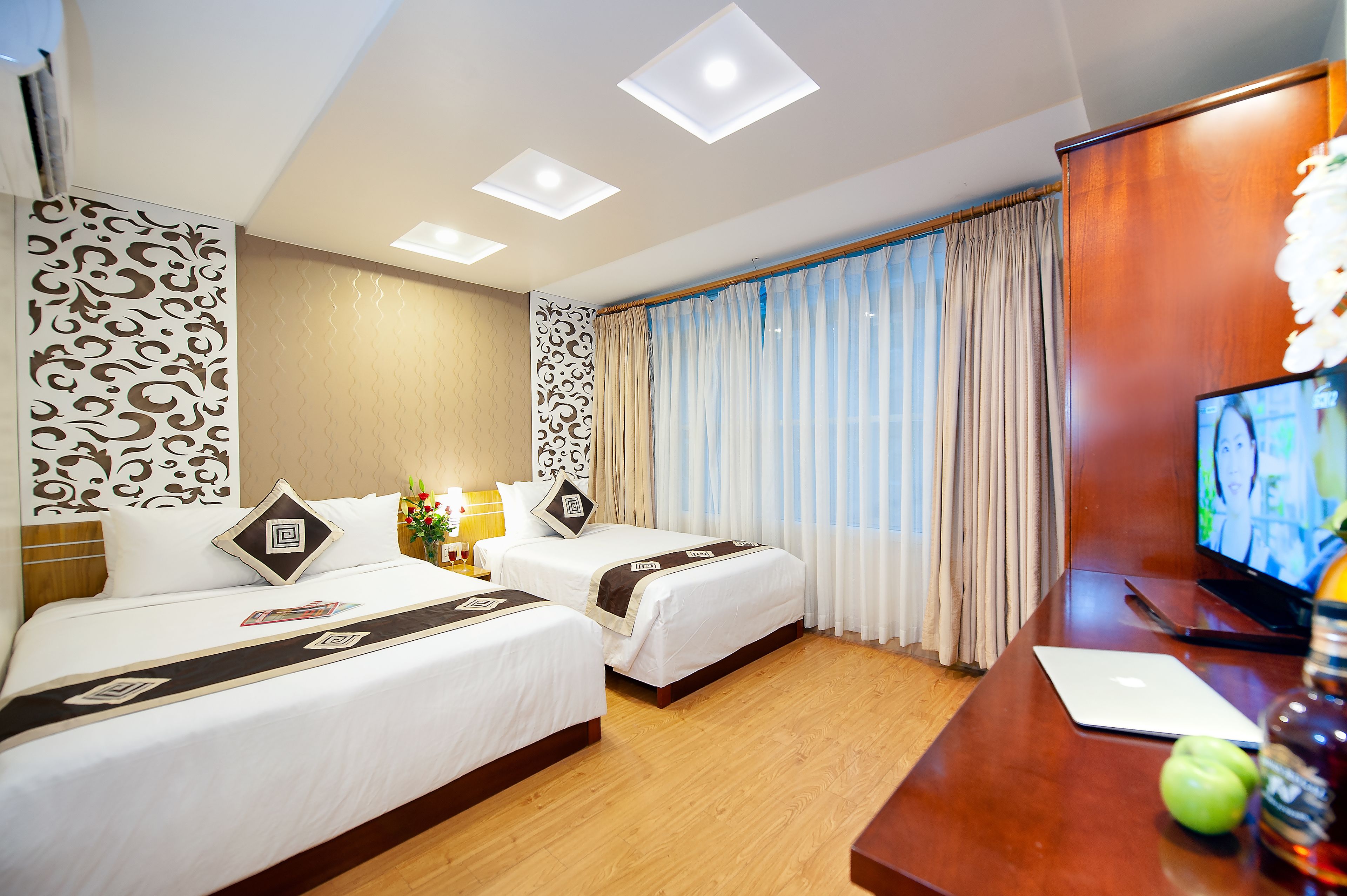 A hotel room with two beds, a TV, a desk with a laptop, and a large window with curtains.