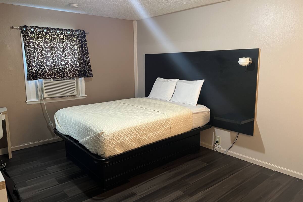 Une chambre à coucher avec un lit, une fenêtre avec des rideaux, un luminaire fixé au mur et une prise de courant.