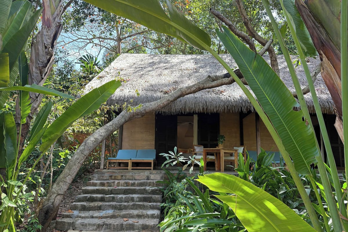 A thatched-roof hut with a wooden bench and a table, surrounded by lush greenery.