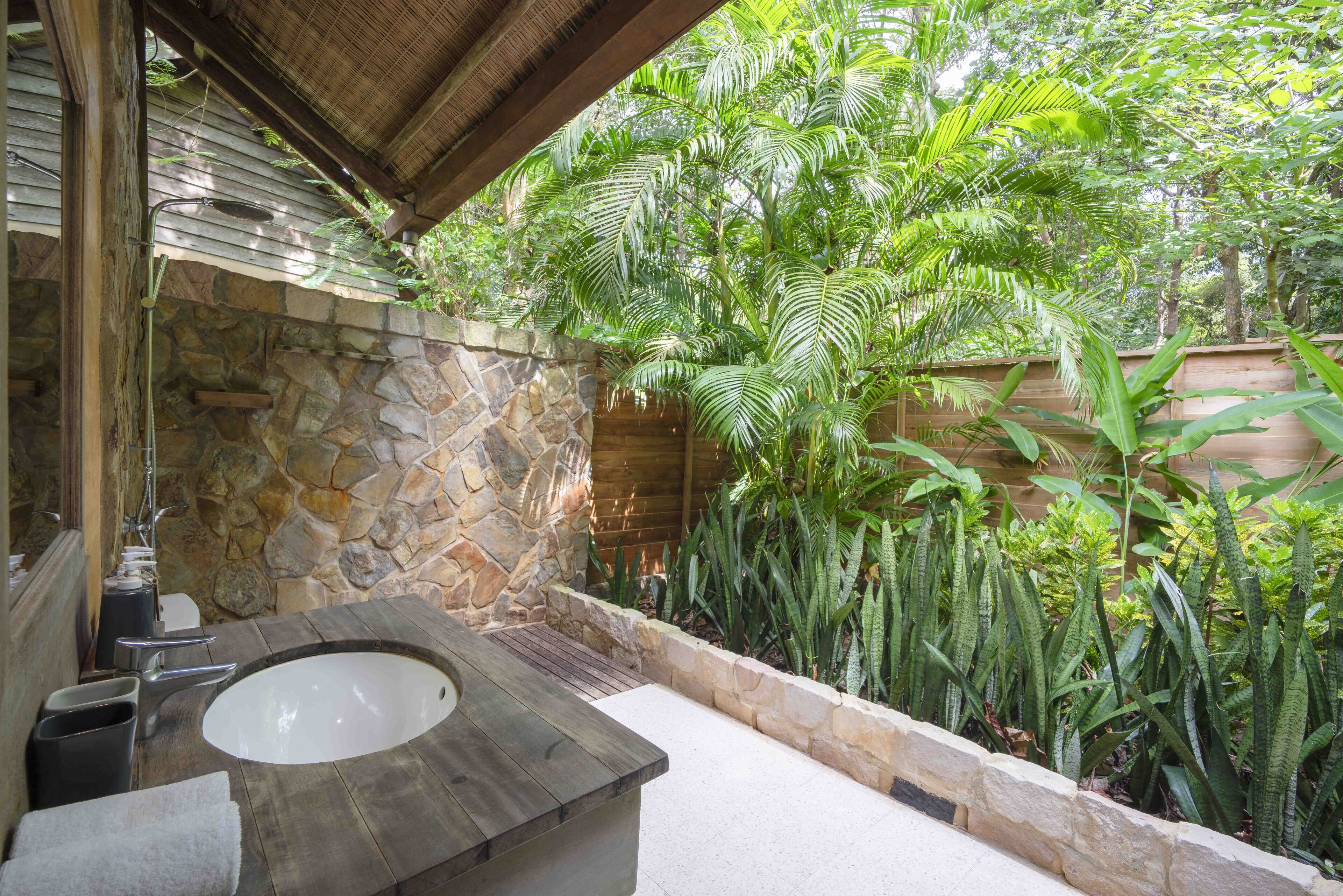 A bathroom with a stone wall, a wooden bathtub, and a variety of tropical plants.