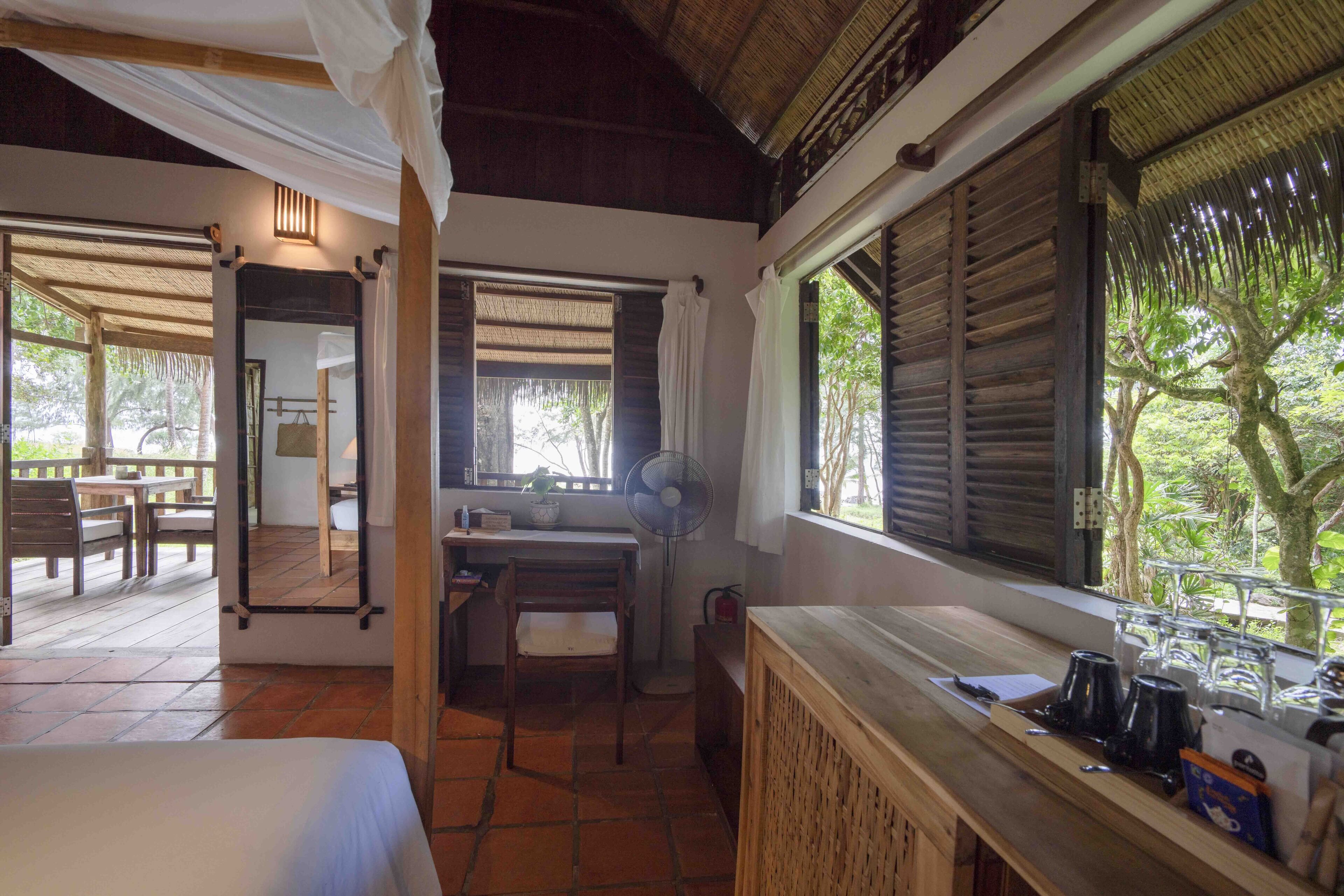 A room with a bed, a desk, a chair, a fan, and a window with a view of trees.