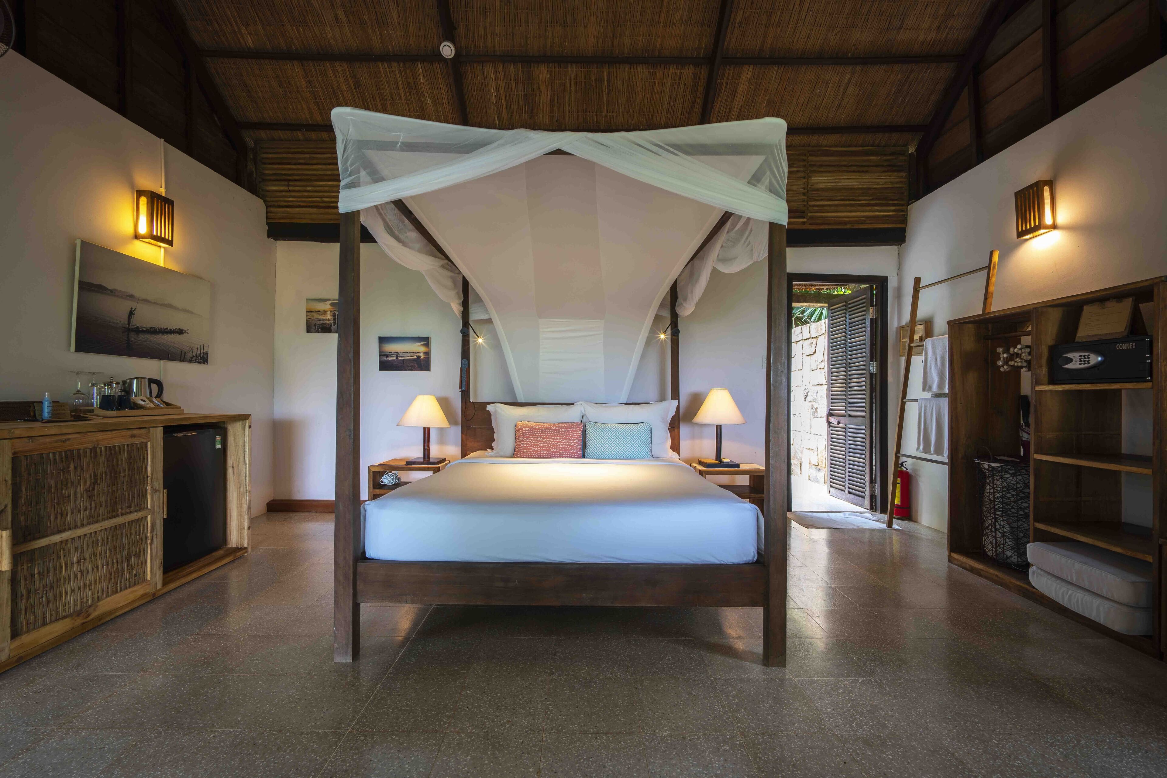 Una cama grande con dosel, lámparas de noche, un aparador de madera y un estante pequeño con un libro y un adorno.
