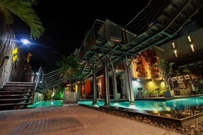 Vista noturna de um hotel moderno com piscina, área de estar externa e escadaria que leva à entrada.