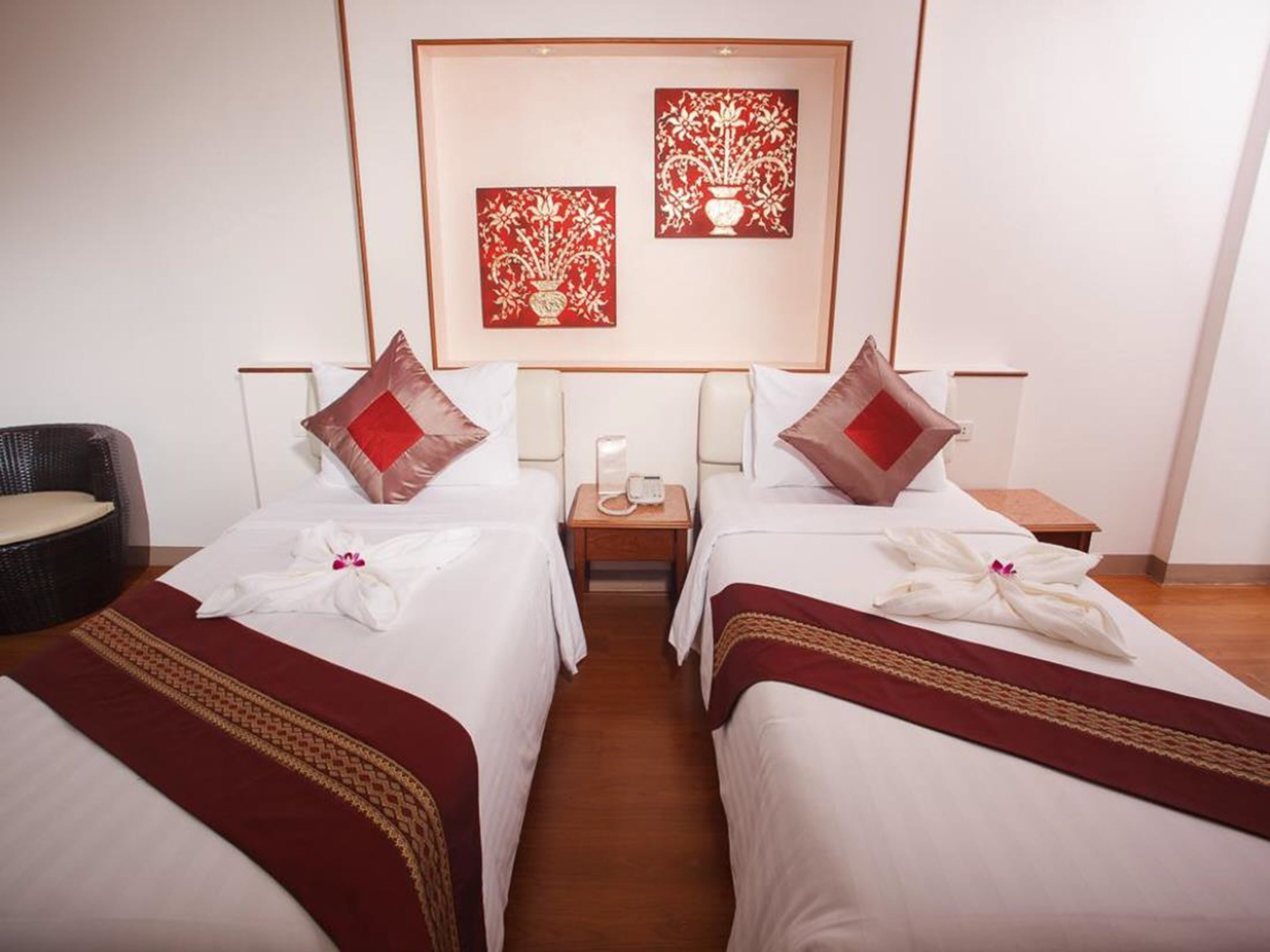 A hotel room with two beds, each with a red pillow and a white towel. There is a nightstand with a phone and a lamp between the beds. The room has a wooden floor and a wicker chair.