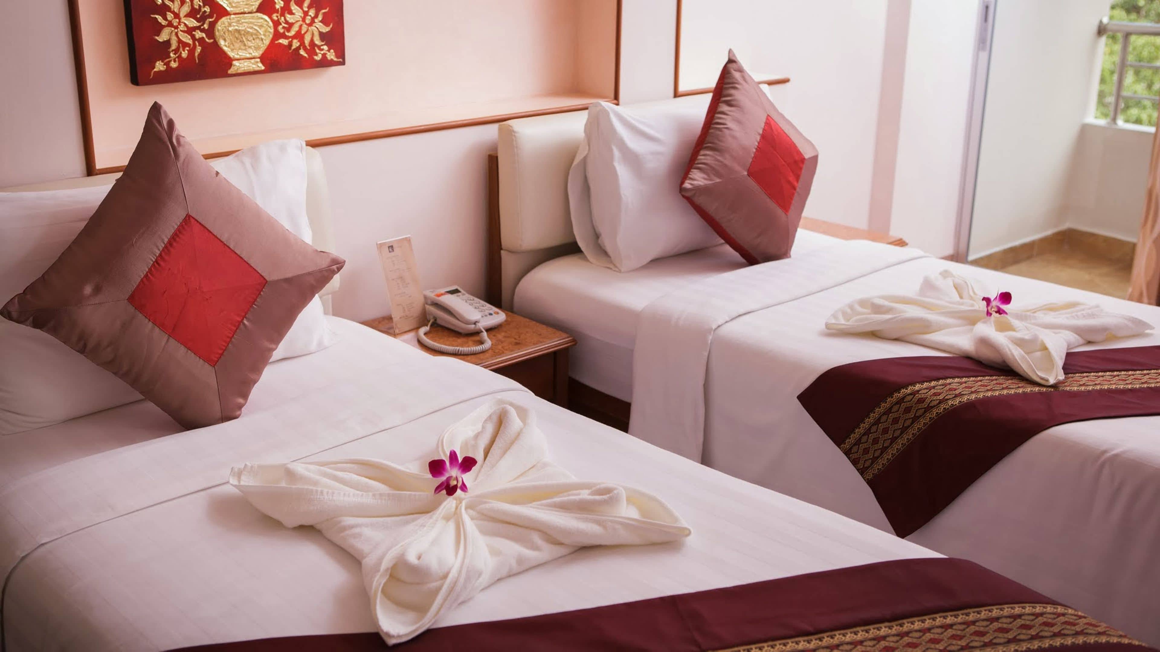 Dos camas de hotel con ropa blanca, almohadas rojas y marrones, y toallas dispuestas en un diseño floral.