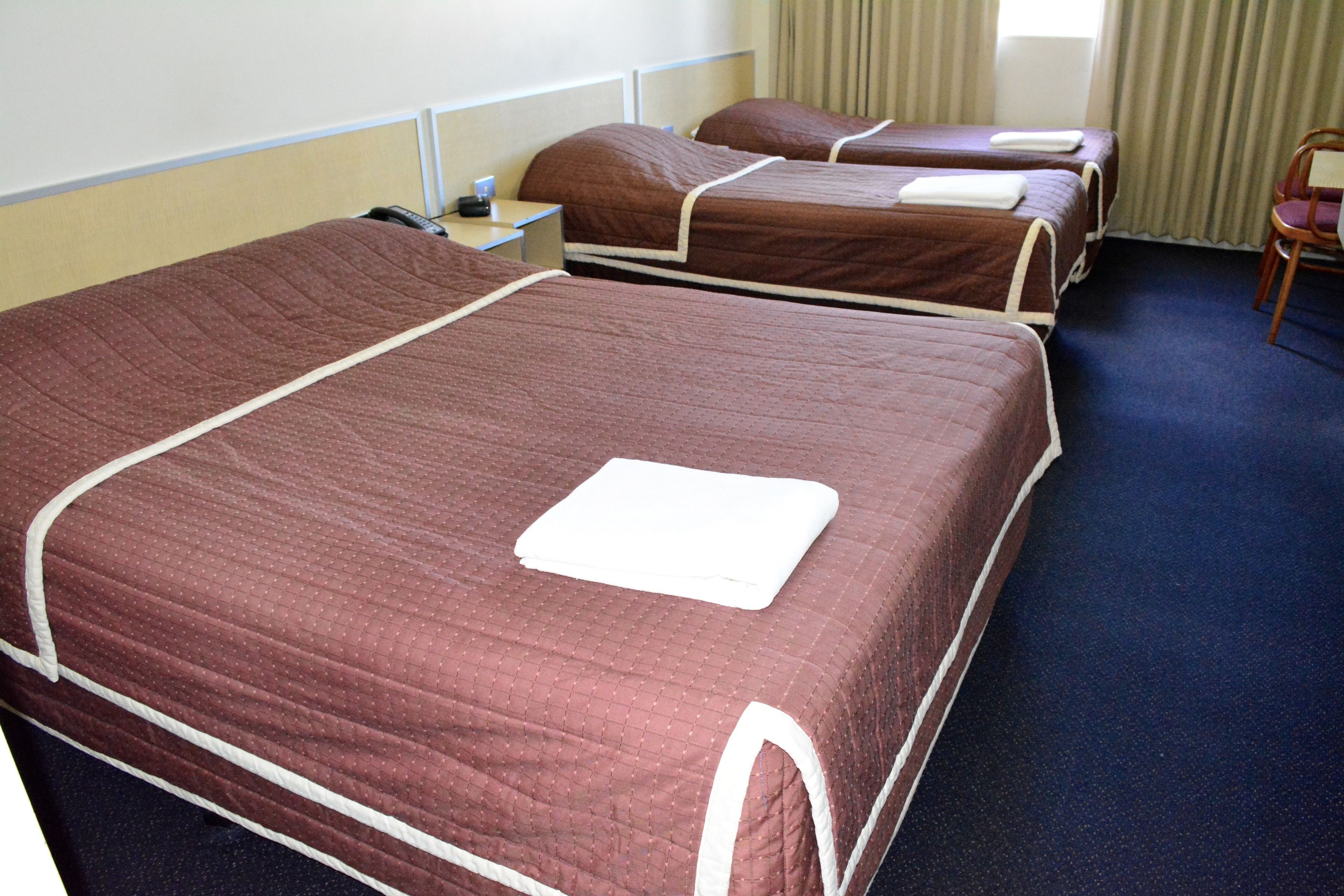 A hotel room with three beds, each with a brown quilted bedspread and white trim. There is a small bedside table with a lamp and a phone.