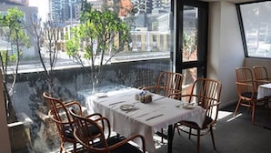Un comedor con una mesa preparada para cuatro personas, rodeado de sillas de mimbre, con vista a un paisaje urbano a través de un gran ventanal.
