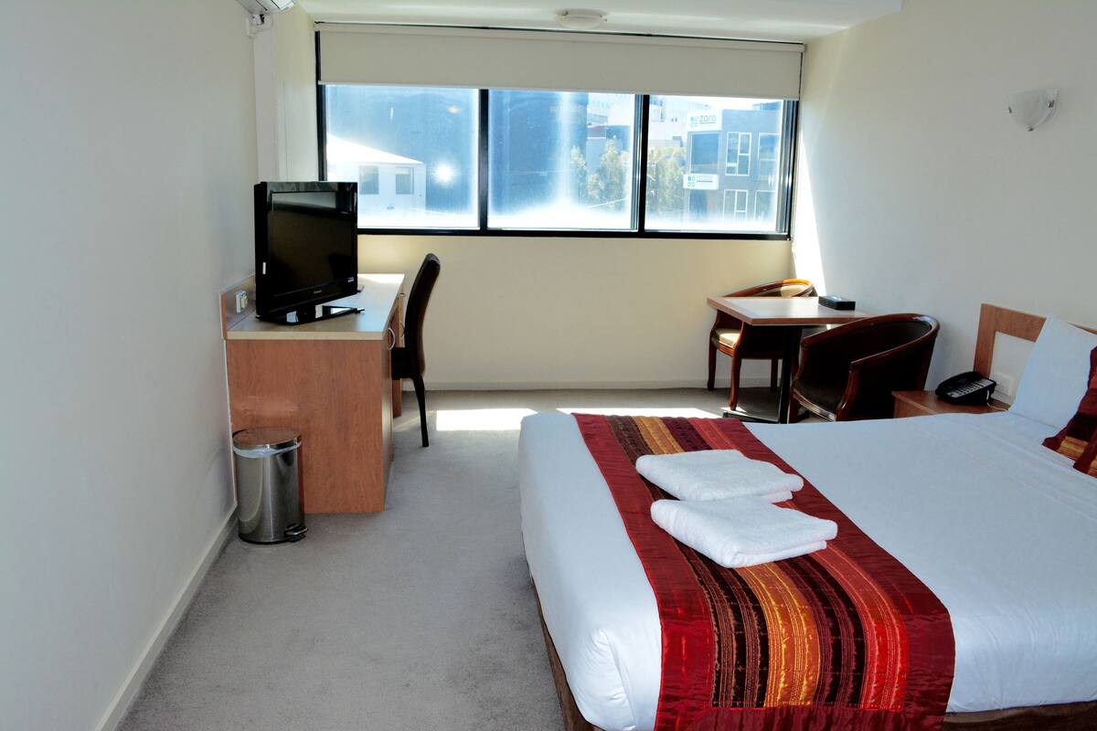 A hotel room with a bed, a desk with a television, a chair, a small table, and a window with a view of buildings.