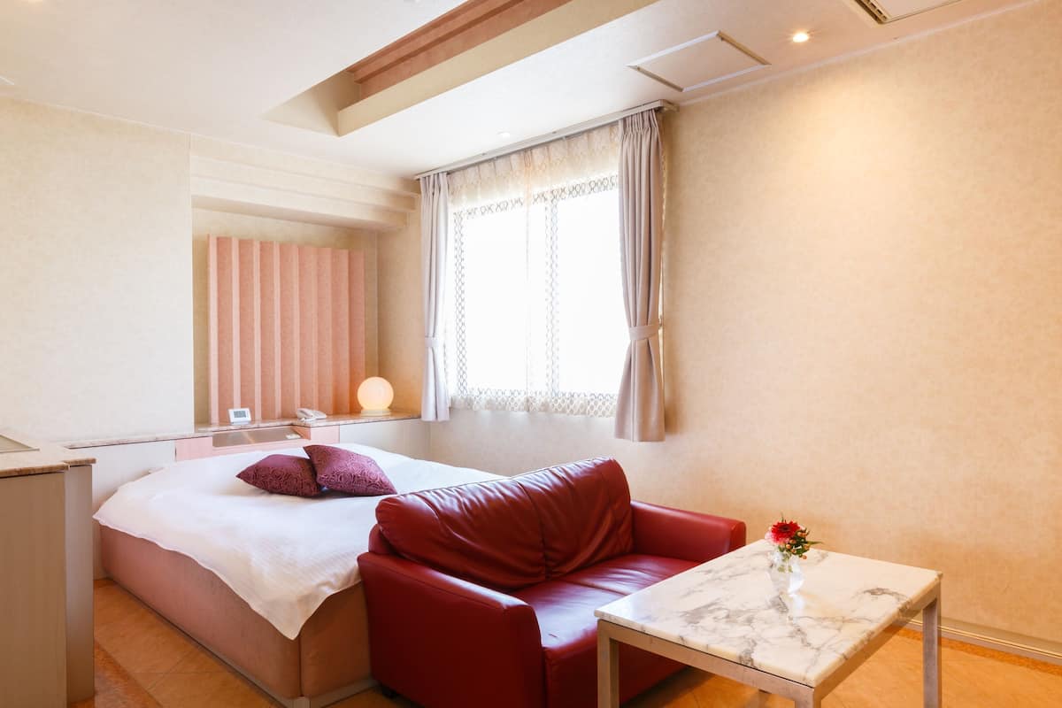 A modern hotel room with a bed, a red leather sofa, a small table, and a window with curtains.