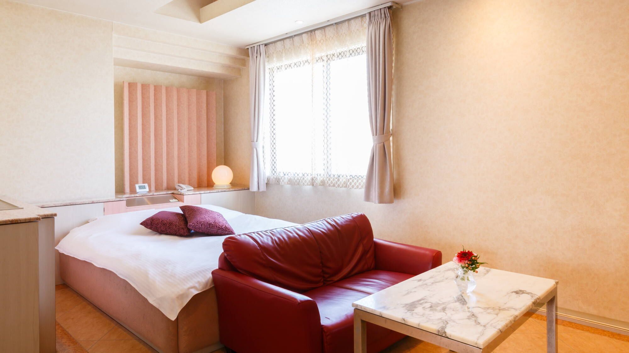 A hotel room with a bed, a red leather sofa, a small table, and a window with curtains.
