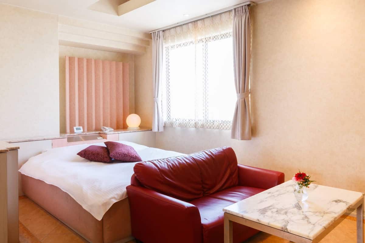 A hotel room with a bed, a red leather sofa, a small table, and a window with curtains.