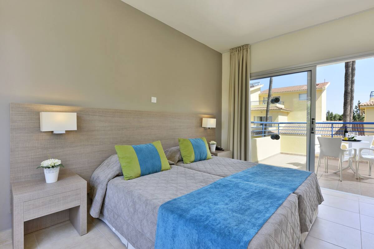 Une chambre d’hôtel comprenant un lit, deux tables de chevet, une petite table, une plante en pot et un balcon avec des chaises et une table.