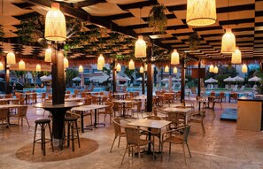 An outdoor dining area with wicker furniture and hanging lamps, overlooking a pool. - Makronisos Holiday Village (Ayia Napa)