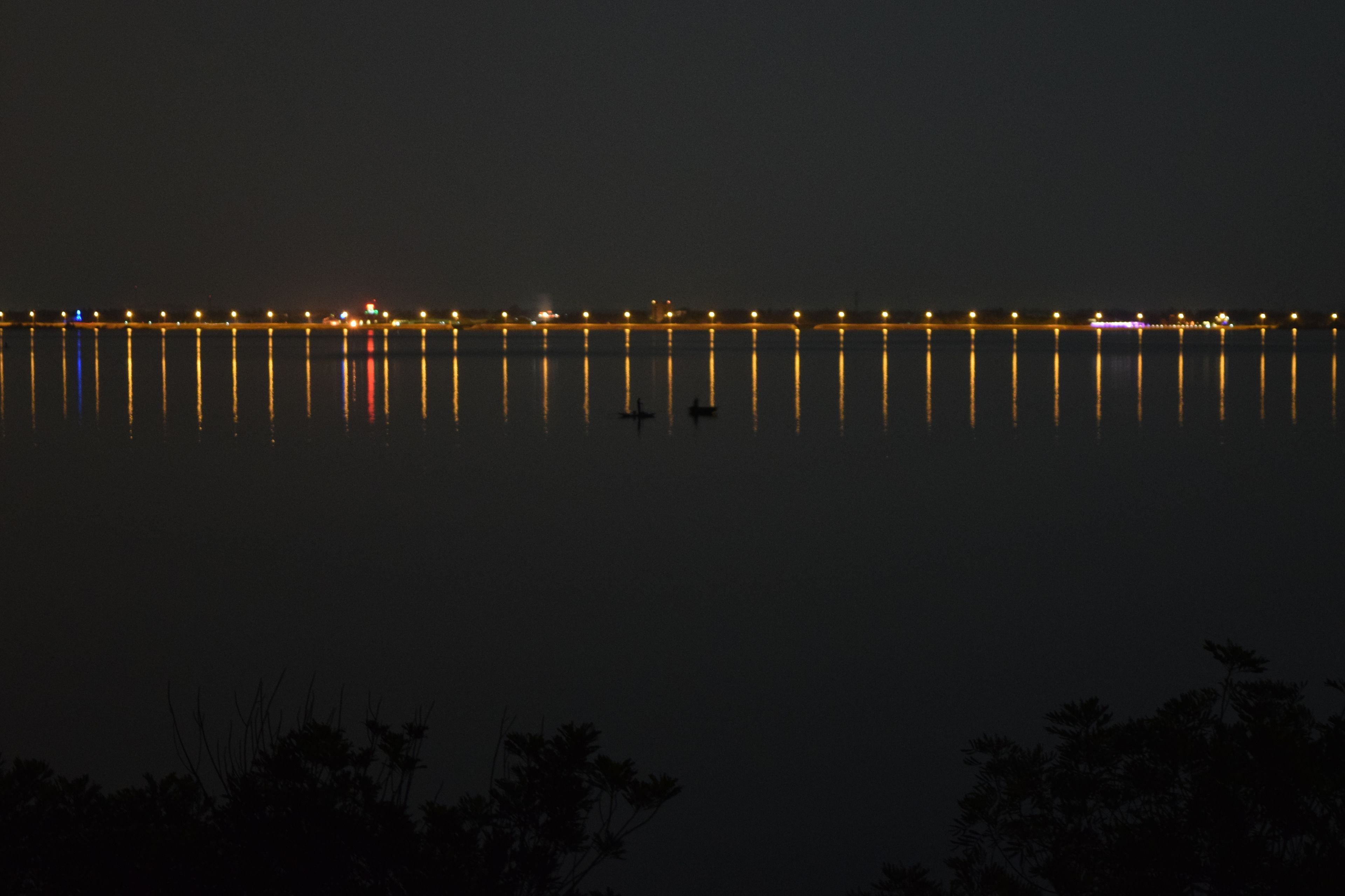 Une vue nocturne d’une étendue d’eau calme, avec un pont et des lumières lointaines qui se reflètent à la surface.