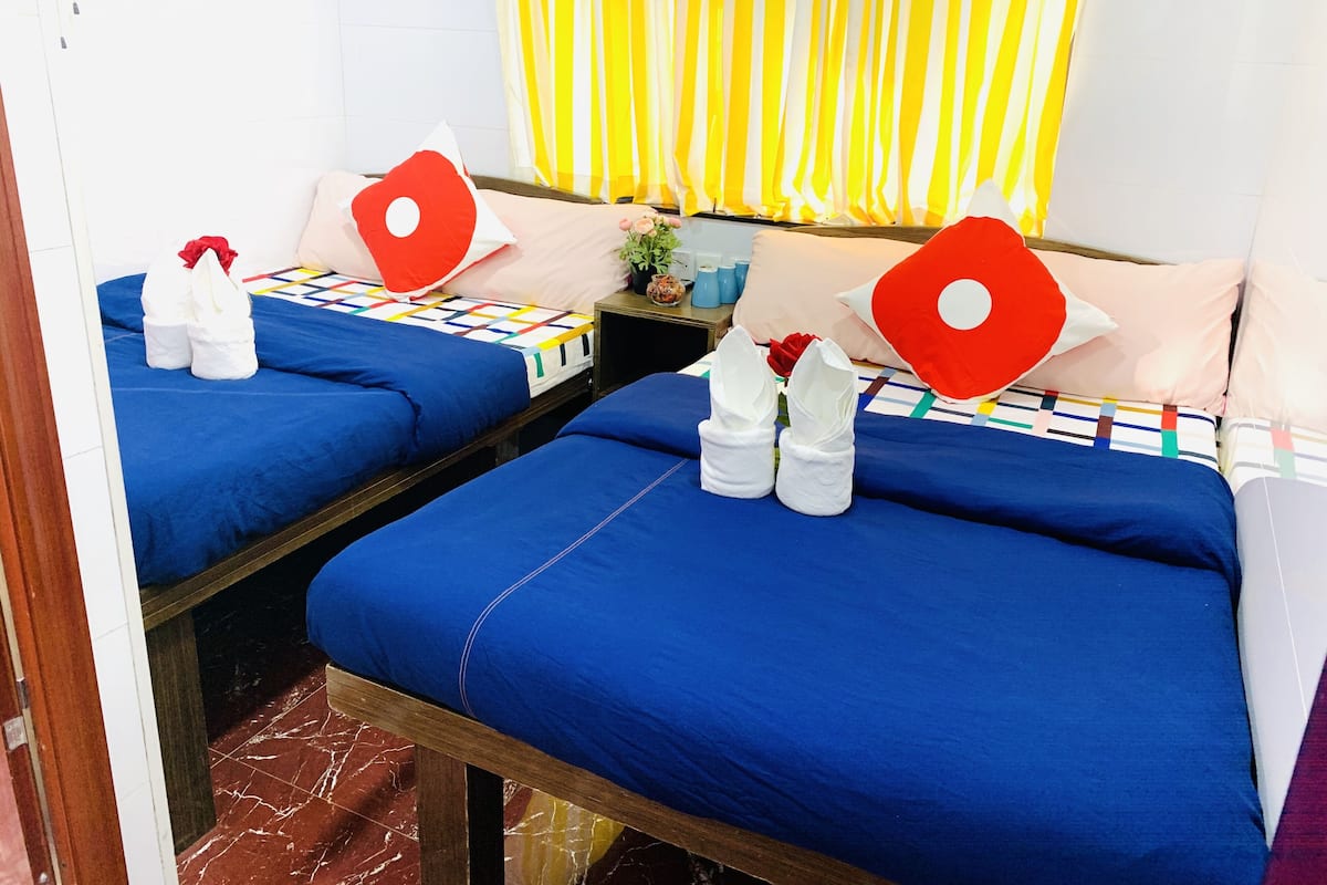 Quarto de hotel com duas camas, cada uma com roupa de cama azul e almofadas vermelhas. Há toalhas brancas nas camas, e uma pequena mesa com um vaso de flores.