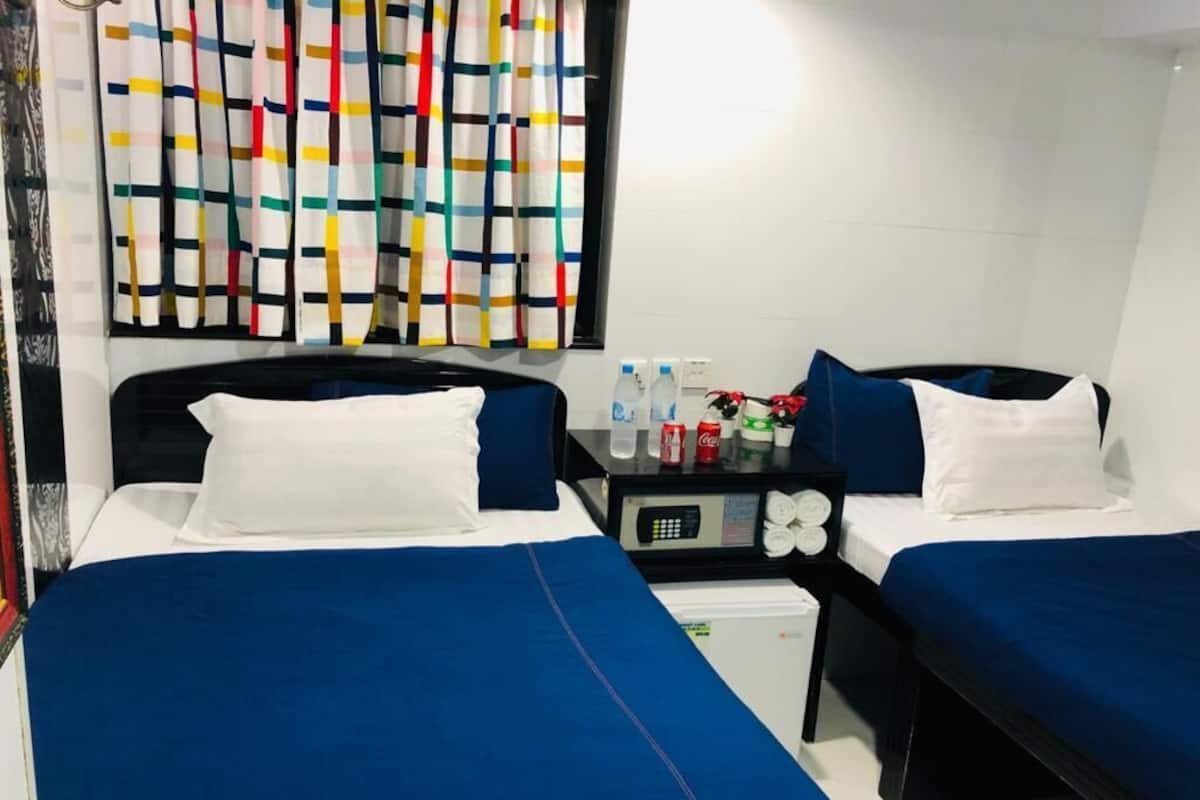 Quarto de hotel com duas camas, cada uma com roupa de cama azul e almofadas brancas. Há uma pequena mesa com um painel de controle e uma geladeira. Uma cortina colorida é visível ao fundo.
