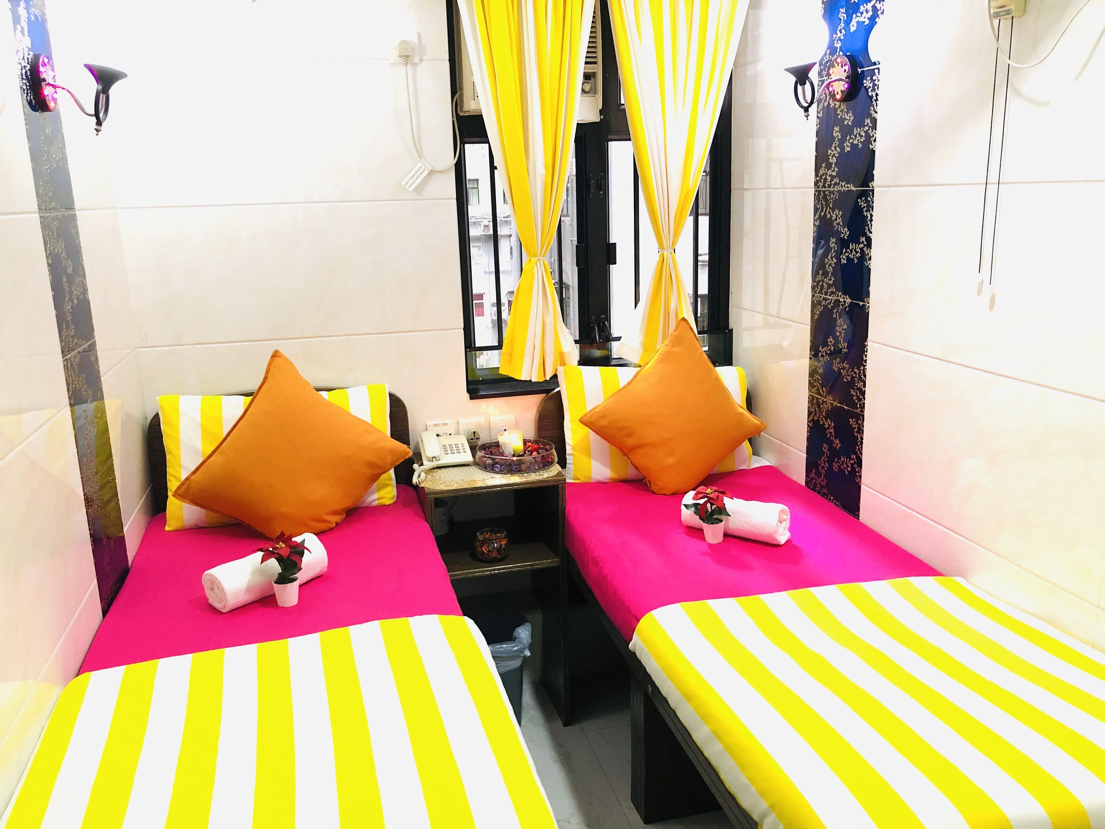 A hotel room with two beds, each with a striped bedspread, pillows, a bedside table with a phone and a small tray of fruit, and a window with yellow curtains.