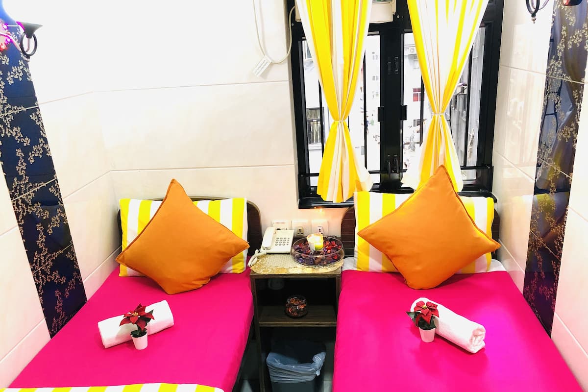 Quarto de hotel pequeno com duas camas, cada uma com colcha listrada em rosa e amarelo, e almofadas a combinar. Há uma mesinha entre as camas com um telefone e uma tigela de frutas.