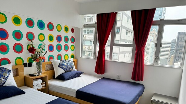A hotel room with two beds, a colorful wall decoration, a small table with a vase, and a view of the city through large windows. - Ashoka Hostel (Kowloon)