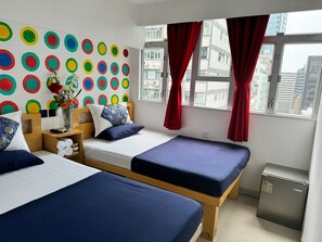 A hotel room with two beds, a colorful wall decoration, a small table with a vase, and a view of the city through large windows. - Ashoka Hostel (Kowloon)