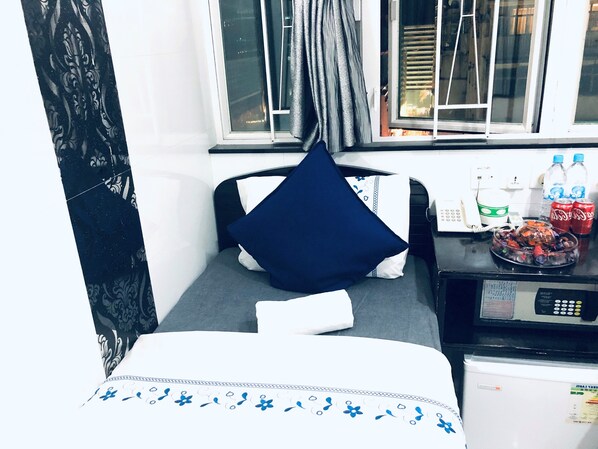 Una habitación de hotel pequeña con una cama, un escritorio con teléfono y un frutero, una ventana con cortinas y un refrigerador.