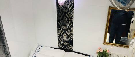 Un lit bien fait, avec du linge de lit blanc et des motifs floraux bleus, une table de chevet avec un téléphone et un petit objet décoratif, et un ventilateur fixé au mur.