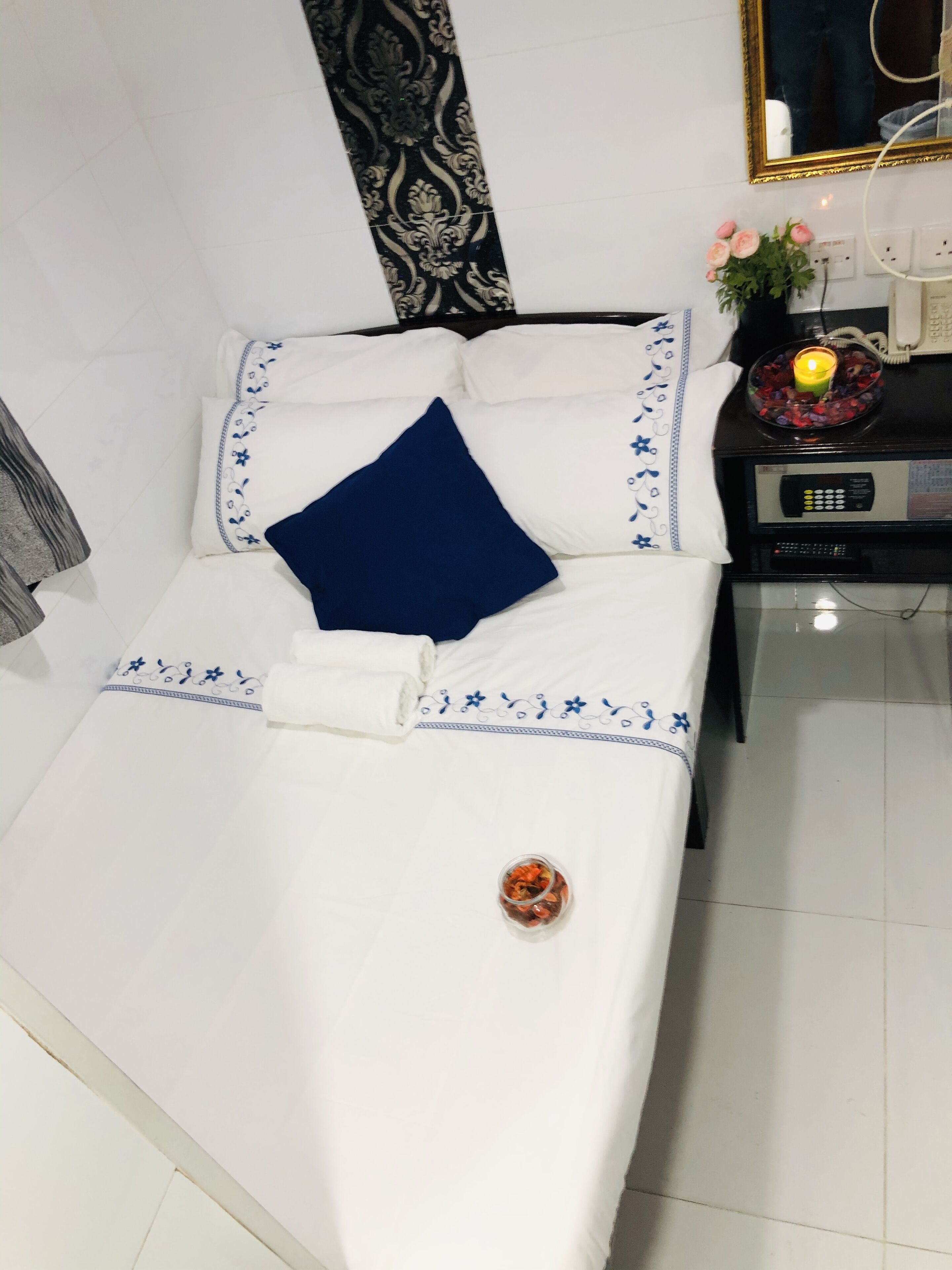 Una cama bien hecha con ropa blanca y detalles azules, una mesita de noche con un teléfono y un adorno, y una pared con un diseño decorativo.