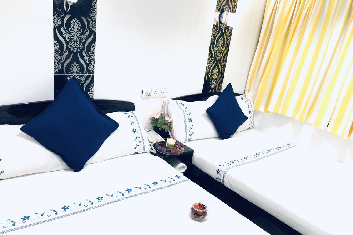 Een hotelkamer met twee bedden, elk voorzien van wit beddengoed en blauwe kussens, een klein nachtkastje met een kaars en fruit, en kamergordijnen met een patroon.
