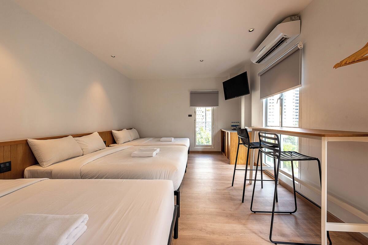 A hotel room with three beds, a desk with a chair, a TV mounted on the wall, and a window providing a view of the outside.