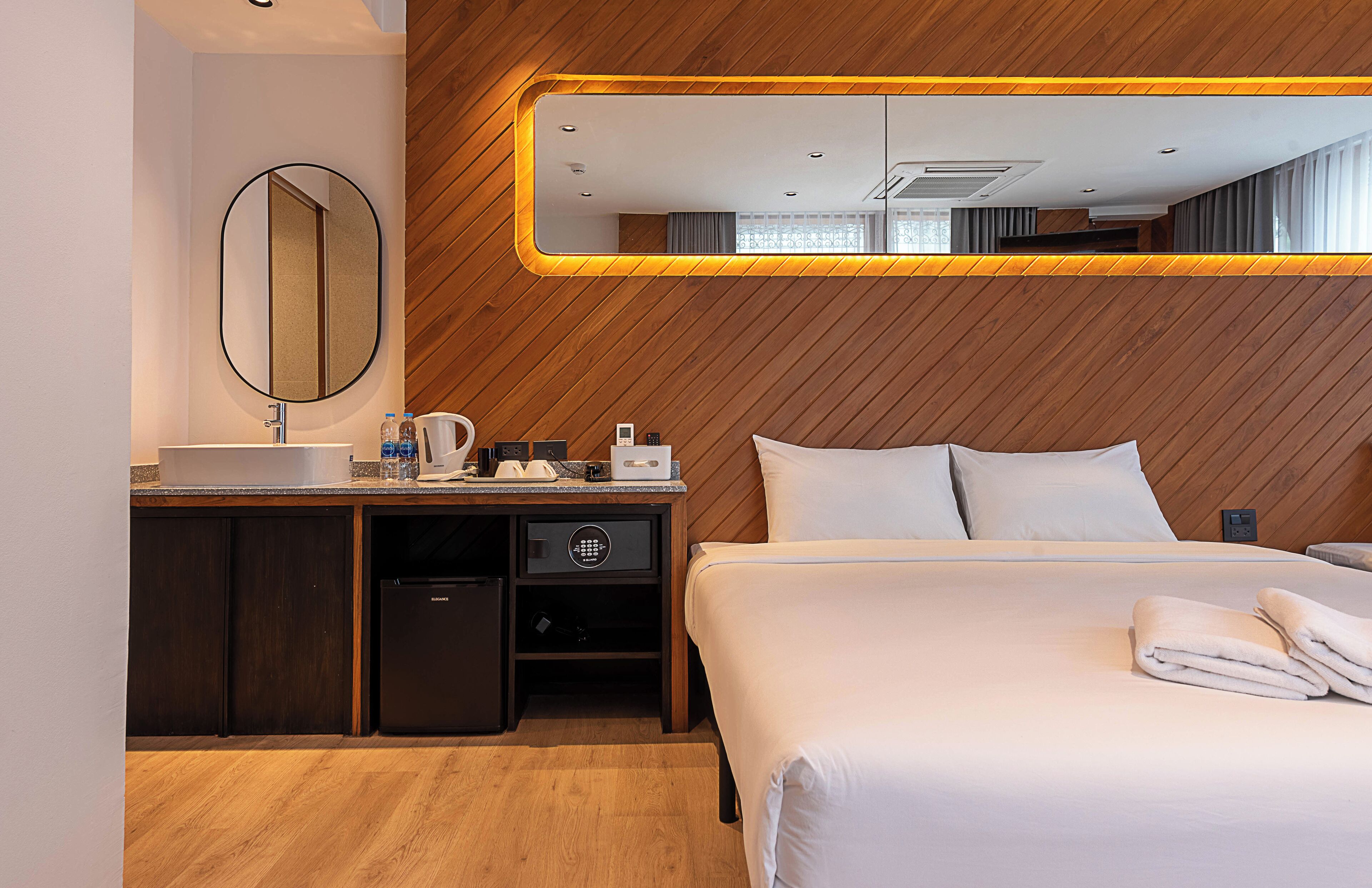 A hotel room with a wooden wall, a large mirror, a bed with white linens, a bedside table with a lamp, and a built-in desk with a sink and a coffee machine.