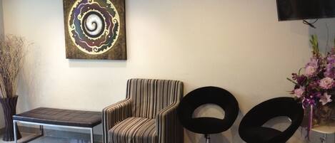 Un vestíbulo con un sillón a rayas, una silla negra y un banco. Hay un televisor montado en la pared y una pieza decorativa sobre el sillón.