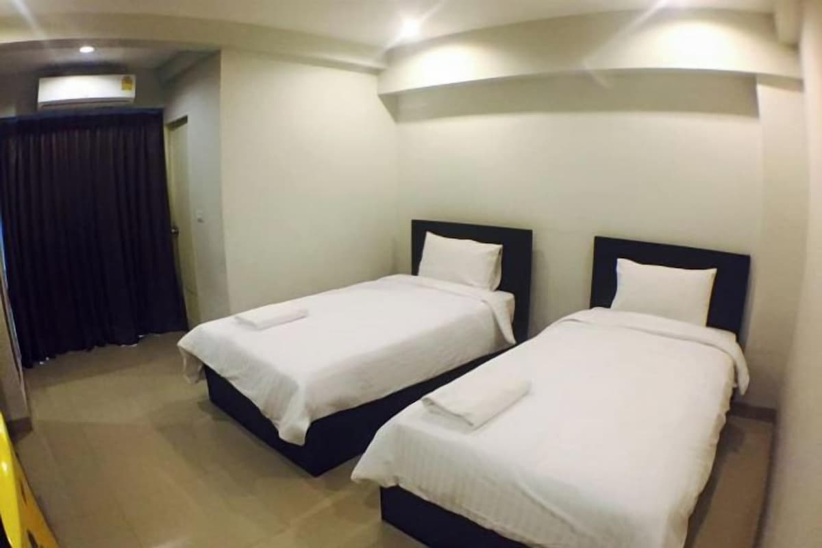 A hotel room with two single beds, a dark-colored headboard, a white wall, and a ceiling-mounted air conditioner.