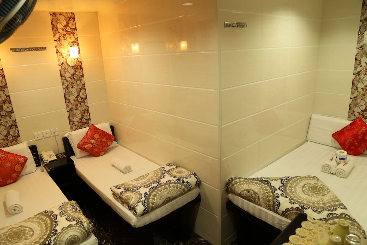 A hotel room with two beds, each with patterned bedding and red pillows. There is a small table with a phone and a bottle of lotion. The walls are tiled, and there is a mirror above the beds.