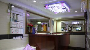 A hotel reception area with a wooden reception desk, a large mirror, and a white sofa with a purple pillow. - Australian Guest House (Kowloon)