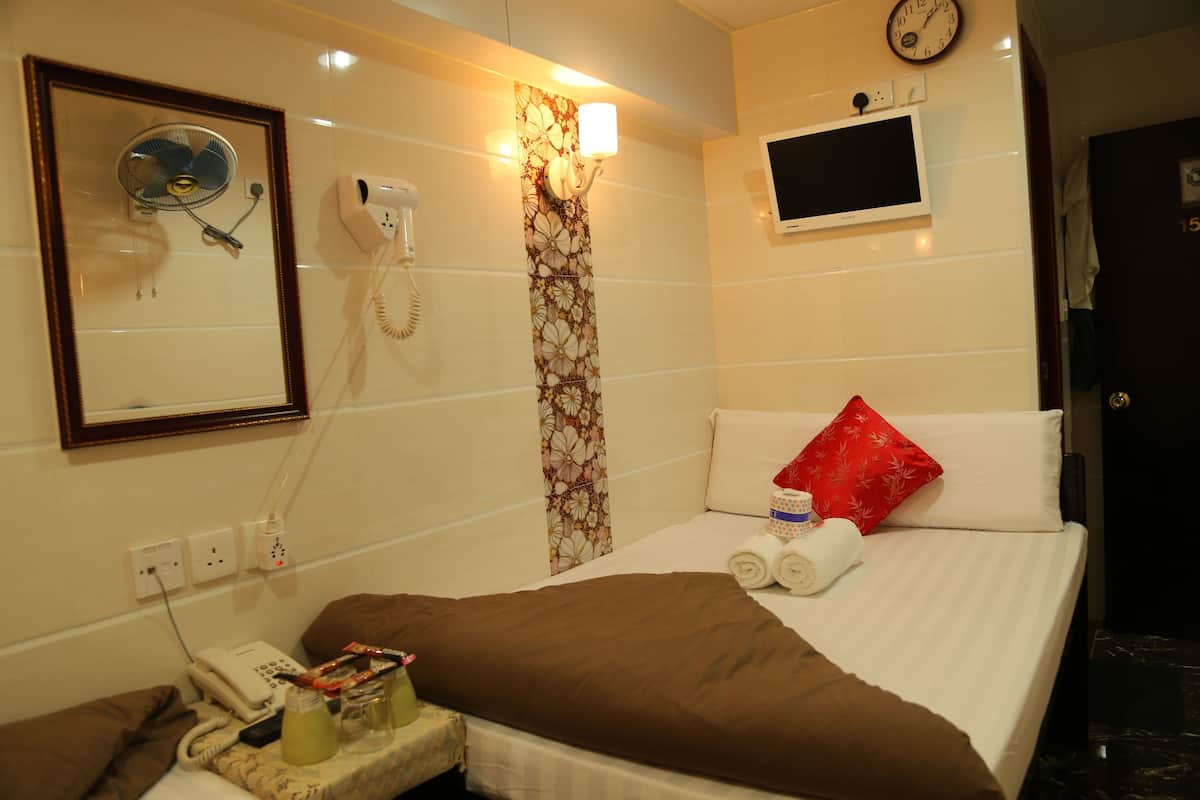 A hotel room with a bed, a bedside table with a phone and a fan, a clock, and a TV on the wall.