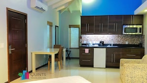 Una cocina compacta con microondas, cafetera y un refrigerador pequeño. Cuenta con una mesa de comedor con sillas y una ventana con persianas.
