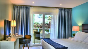 A hotel room with a bed, desk, chair, and a view of a balcony with plants.
