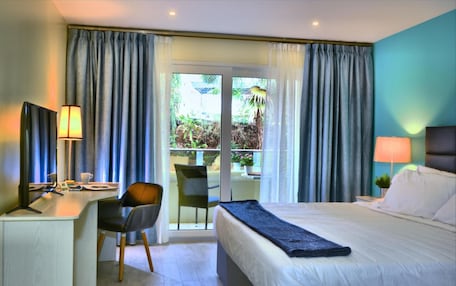 A hotel room with a bed, desk, chair, and a view of a balcony with plants.